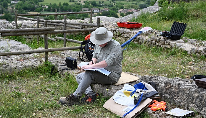 A photo of student Charles Jackson, in Italy working at the Villa of Titus field school. He is seated on a stone wall, writing in a notebook.