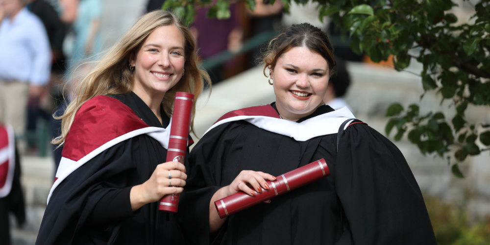 Two graduates hold diplomas while standing outdoors