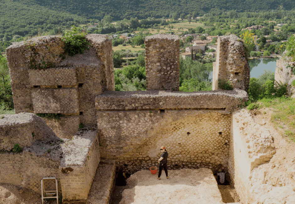 An overhead shot of the archaelogical site in Italy, with green mountains and water shown behind the ancient building. A student stands in front of a wall looking up toward the camera, they are small in comparison to the building.