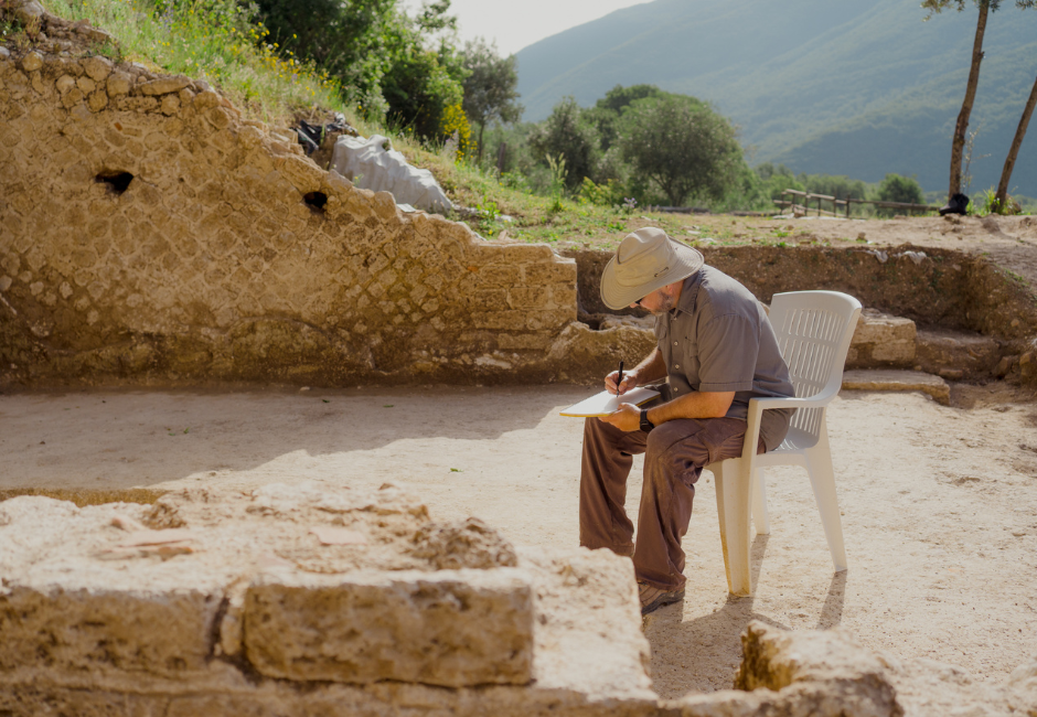 Myles McCallum sits in a chair at an outdoor archelogical site in Italy reviewing documents