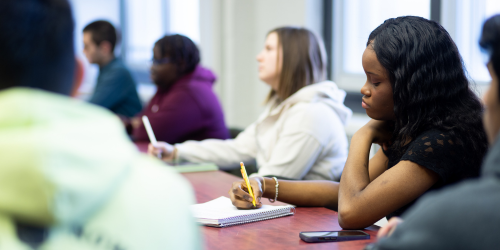 Students sit in a classroom taking notes with pen and paper, they sit facing an unseen lecturer