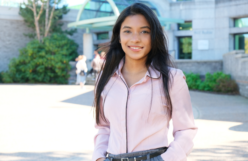 A student with olive skin and long brown hair stands in front of the Sobey School building. She wears a pink button up shirt and dress pants.