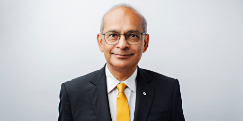 Vivek is a man with short grey hair and round glasses. He wears a dark suit with a bright yellow tie.