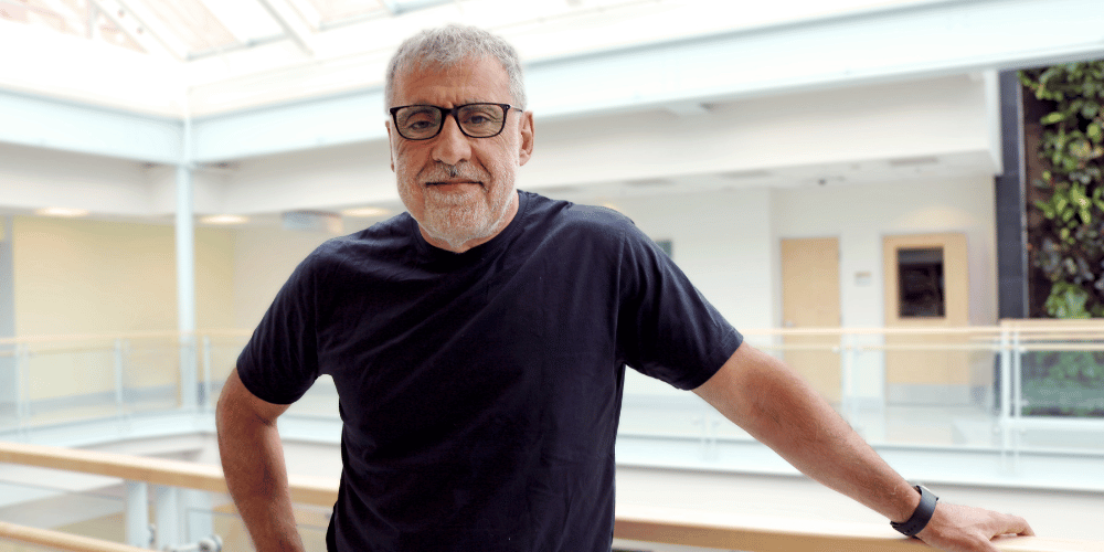 Yigit, a man with tan skin and short grey hair and beard, stands facing the camera. He wears black framed glasses and a black tshirt.