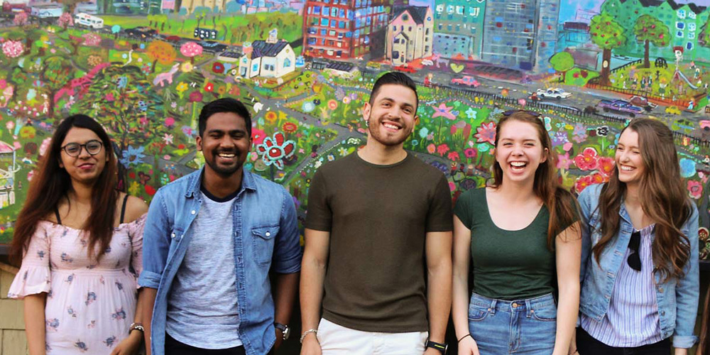 Five smiling students lined up against a wall covered in a painting of a streets and houses.