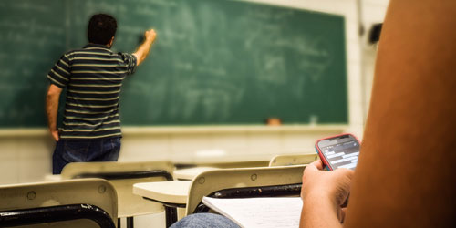 student at chalkboard, another seated with mobile device in hand