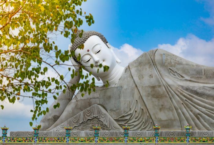 Photo of a reclining Buddha statue under a blue sky with green leaves in Wat Pătum Wôngsa Som Rông, Phường 5, Sóc Trăng, Soc Trang, Vietnam